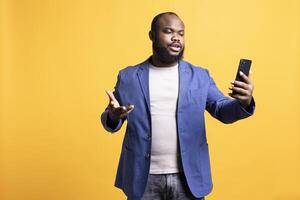 Annoyed man having unpleasant conversation with friends during teleconference meeting using smartphone, studio background. Hostile person having uncivil chat with mates during online call photo