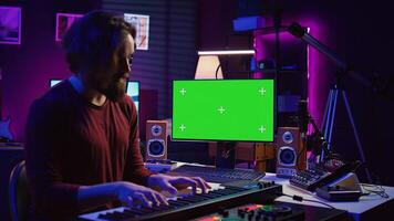 Artist learning to play at electronic piano keyboard in home studio, watching tutorial online with greenscreen display. Music producer practices playing on midi controller synthesizer. Camera B. photo