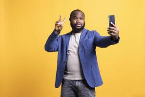 Man using smartphone to take selfies, doing victory hand sign. African american person taking photos using phone selfie camera, showing peace symbol gesturing, studio background