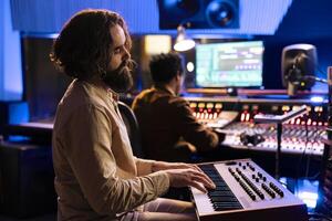 Young artist songwriter creating a new song with piano keys in studio, collaborating with audio engineer in control room. Musician singer composing music with midi controller electronic keyboard. photo