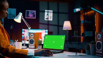 African american girl working with device showing greenscreen layout, sitting at desk and looking at chromakey display. Young woman using laptop with copyspace mockup template. Camera B. photo