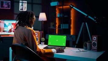 African american young woman looks at laptop with greenscreen, sitting at her home desk and preparing to attend online university lessons. Girl using pc with isolated mockup layout. Camera B. photo