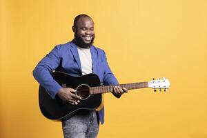 Smiling singer holding guitar, performing blues tunes during concert isolated over yellow studio background. Merry musician performing composition on stringed musical instrument photo