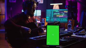 Composer performing acoustical guitar in his home studio, playing and practicing on strings. Musician learning multiple songs on instrument, uses tablet to run greenscreen display. Camera B. photo