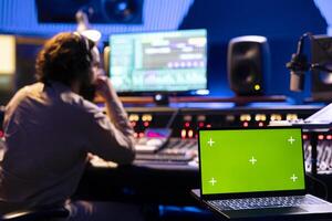 Male technician working on editing tracks next to greenscreen on laptop, operates buttons and knobs in control room. Audio engineer using faders and other technical equipment, mix and master. photo