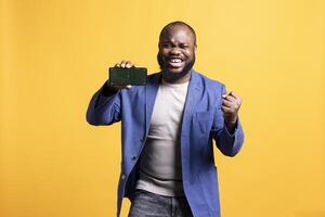 African american man excitedly celebrating while presenting isolated screen phone, studio background. Ecstatic BIPOC person holding copy space mockup cellphone used for advertising brands photo