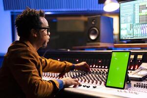 African american music producer recording and editing tracks in studio, operates panel board next to mockup on device. Sound designer mixing and mastering tunes on audio console, stereo gear. photo