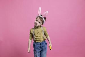 Small positive boy smiling in studio while he wears fluffy bunny ears to celebrate easter holiday festivity. Joyful happy little child standing against pink background, plays with a cute toy. photo