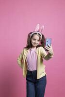 positivo linda Chica de escuela tomando imágenes con su móvil teléfono en estudio, sonriente a cámara y posando con confianza en contra rosado antecedentes. alegre joven niño toma fotos para Pascua de Resurrección festividad.