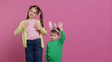 Joyful little kids waving in front of camera during easter holiday, smiling and wearing bunny ears. Brother and sister toddlers greeting someone in studio, adorable happy children. Camera A. photo