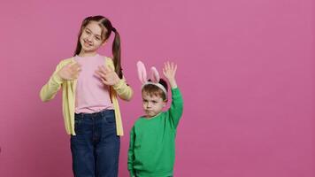 alegre pequeño niños ondulación en frente de cámara durante Pascua de Resurrección día festivo, sonriente y vistiendo conejito orejas. hermano y hermana niños pequeños saludo alguien en estudio, adorable contento niños. cámara una. foto