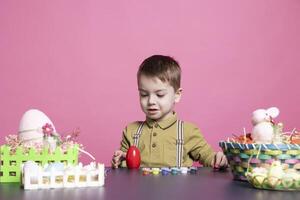 Lovely small boy decorating eggs with watercolors and imprints while making gorgeous ornaments for Easter Sunday event. Adorable youngster enjoys coloring with artistic supplies in the studio. photo