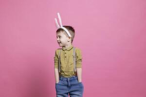 Cheerful young kid smiling and wearing fluffy bunny ears on camera, feeling joyful about easter festivity celebration and standing against pink background. Adorable small boy with colorful outfit. photo