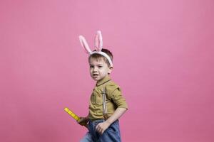 joven adorable niño jugando con un el plastico juguete en frente de cámara, vistiendo conejito orejas durante Pascua de Resurrección fiesta celebracion. alegre sonriente pequeño chico disfrutando primavera evento en contra rosado fondo. foto