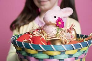 Little kid showing colorful easter decorations and painted eggs in a basket, holding spring festive arrangement in pink studio. Young small girl smiling on camera with lovely toys. Close up. photo