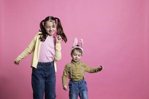 Cute young children dancing around in front of camera, feeling happy and curious about easter celebration or gifts. Brother and sister enjoying spring holiday, little boy wearing bunny ears. photo