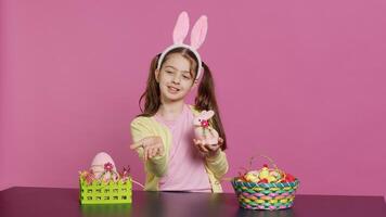 Young schoolgirl blowing air kisses in front of camera while she creates festive lovely arrangements on stuffed rabbit toy. Cheerful young child being excited about easter celebration. Camera A. photo