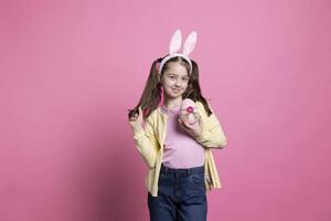 Little young girl with fluffy bunny ears shows a pink easter egg in front of camera, holding her handmade festive spring ornaments over studio background. Small child with arrangements. photo