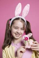 alegre bonito niñito con conejito orejas y coletas participación un rosado Conejo juguete, posando con confianza terminado rosado antecedentes. joven niña sensación optimista acerca de Pascua de Resurrección celebracion, vistoso decoración. foto