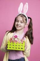 Small joyful girl with fluffy ears showing cute painted basket filled with eggs in front of camera, posing with festive decorations. Young little child being enthusiastic about easter celebrations. photo