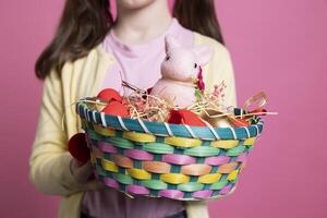 Small child presenting easter decorations and toys in a basket, holding colorful spring festive arrangements in front of camera. Young positive girl smiling in studio with adorable decor. Close up. photo