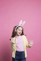 Little child acting excited and cheerful about easter festivity, posing with confidence against pink background. Young small kid wearing bunny ears and pigtails, feeling cheerful and carefree. photo