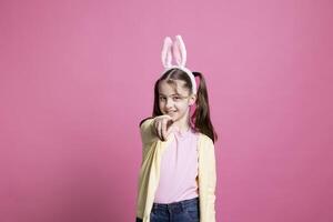 Young schoolgirl with pigtails dancing around in pink studio, pointing at something in front of the camera and having fun. Joyful small child feeling confident with bunny ears for easter. photo
