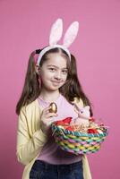 sonriente linda pequeño niña demostración vistoso huevos y Conejo juguete en un acuerdo, posando con confianza en rosado estudio. joven niño con conejito orejas participación un cesta con encantador decoraciones foto