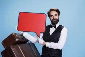 Hotel worker showing ad with speech bubble, holding empty isolated red billboard icon. Elegant doorkeeper presents blank copyspace cardboard sign on camera against blue background. photo