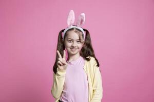 sonriente pequeño niño demostración paz firmar en frente de cámara, sensación emocionado acerca de regalos y Pascua de Resurrección festividad. joven niña con conejito orejas posando en estudio, siendo contento y positivo acerca de primavera. foto