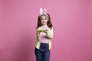 adorable pequeño niña demostración un cesta lleno con Pascua de Resurrección huevos y un polluelo, posando con confianza en frente de cámara. alegre pequeño niño participación primavera decoraciones, coletas y conejito orejas. foto