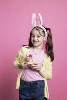 alegre pequeño niño posando con un rosado Conejo juguete en cámara, vistiendo mullido conejito orejas y participación Pascua de Resurrección adornos terminado antecedentes. joven niñito con coletas siendo emocionado acerca de festivo evento. foto