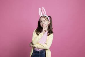 Small cheerful girl acting thoughtful about easter gifts in studio, thinking about holiday celebration with her bunny ears. Sweet young child with pigtails feeling confident in front of camera. photo