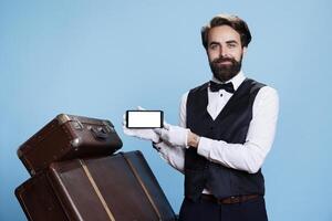 Professional bellman using mobile device with white screen, being dressed in appropriate clothing to represent hospitality industry. Bare copyspace template is highlighted by classy hotel staff. photo
