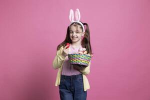 Adorable joyful child holding colorful eggs and a stuffed rabbit on an easter arrangement, posing in pink studio. Young smiling girl with bunny ears holding a basket with lovely spring ornaments. photo