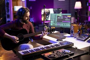 joven cantante jugando acústico guitarra y creando música con electrónico herramientas en hogar estudio, utilizando grajilla software interfaz en ordenador personal. sonido productor grabación melodías y mezcla sonidos en estéreo consola. foto