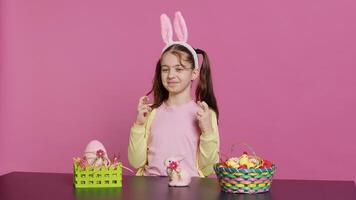 pequeño alegre niño posando con dedos cruzado en estudio, sentado a un mesa lleno con Pascua de Resurrección festivo adornos joven Chica de escuela esperando para suerte y fortuna durante primavera fiesta celebracion. cámara una. foto