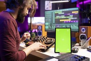 compositor creando bandas sonoras en estéreo engranaje y pantalla verde monitor en móvil dispositivo, mezcla y dominar melodías en hogar estudio. joven artista ajustando volumen niveles en música, grajilla software. foto
