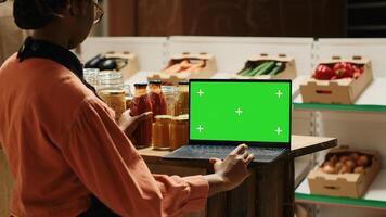 Young vendor looks at greenscreen on laptop while preparing merchandise for customers, using isolated mockup layout on screen. African american seller checking blank chromakey display. Camera 1. photo