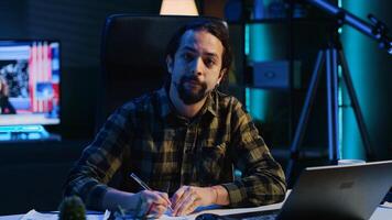 Entrepreneur working at home office desk on laptop, browsing the internet and writing in notebook. Caucasian man using pen, notepad and digital device while teleworking, camera A photo