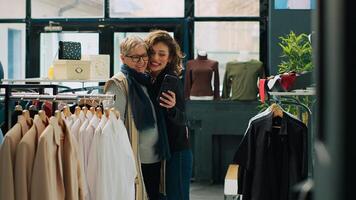 Senior woman and her daughter take photos in department store, enjoying day off shopping for clothes. Smiling customers have fun with selfies in a fashion showroom, commercial activity. Camera A.