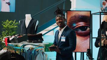 African american worker managing clothes stock on hangers, working on inventory using tablet in shopping center. Male store assistant in a suit preparing fashion collection on display. Camera A. photo