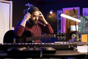 Sound composer operating with electronic setup in home studio, recording and playing acoustic guitar tunes. Musician working with signal processing techniques and audio plug ins at desk. photo