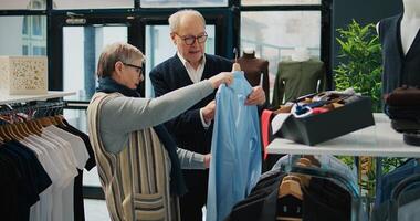 Senior couple shopping for formal outerwear at fashion boutique, looking for trendy elegant clothing items to create new outfits for elderly man. People searching for stylish merchandise. Camera B. photo