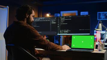 Green screen laptop next to freelancing man doing IT support job from apartment office, coding on PC. Mockup notebook on desk where software engineer writes lines of code on computer at home, camera B photo