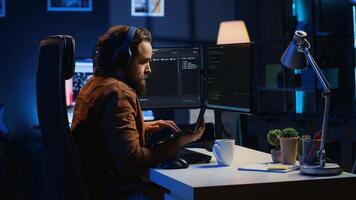 Developer focused on typing complex code in neon lit home office, listening music and developing software application. IT expert wearing headphones, solving coding project tasks on laptop, camera A photo