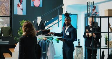 African american worker checking inventory for new clothing line placed on hangers for sale, looking at merchandise on tablet and in store. Retail assistant verifying discount list at mall. Camera B. photo