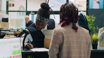 africano americano repartidor recepción solicitado ropa lleno en cajas, llegando a Moda sala de exposición a recoger arriba orden y firmar documentos. mujer empleado preparando artículos a efectivo registro. cámara una. foto