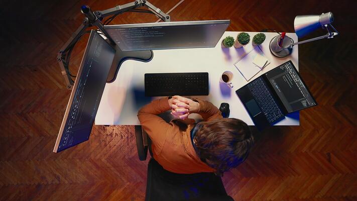 Aerial shot of computer scientist listening music while writing lines of code during remote job shift in home office. IT professional enjoying audio podcast, coding on desktop PC, top down view