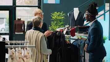 African american worker suggesting shirts from new collection in fashion boutique, discussing with elderly shoppers. Stylist showing clothing items to senior clients, consumerism. Camera A. photo